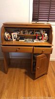 Front view of open rolltop desk filled with various items inside compartments, two drawers below, and cupboard door open showing shelf inside.