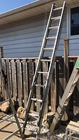 Full view of the extended aluminum ladder leaning against house siding and wooden fence outdoors.