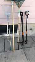 Photo showing 5 yard tools including two types of rakes, squeegee, and two Fiskars weed pullers against a wall