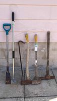 Full set of six hand tools including axe, sledgehammer, ice chippers, adze, and prybar against wall on pavement