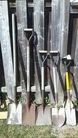 Photo showing five garden tools with wooden handles leaning against a wooden fence, including four shovels and one spade. Handles vary in length; one tool has a yellow fiberglass handle.