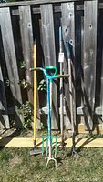Five yard tools including manual tiller, pitchfork, hoes leaning against wooden fence outdoors on grass