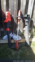 Photo showing the two orange and black Black & Decker garden tools: GrassHog trimmer on the right, and LeafHog with bag on the left leaning against a wooden fence outdoors.