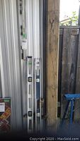 Photo showing two 4-foot aluminum levels leaning against a wall outdoors near a wooden post and a metal shed. The levels have plastic windows displaying bubble vials for leveling, with minor usage marks.