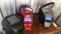 Lot overview showing clear plastic safety visor and three welding helmets placed side by side on a wooden surface.