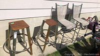 Four stools placed outdoors against an exterior wall: two metal stools with wooden square seats showing rust and wear, and two wrought iron stools with woven seats and wooden armrests.