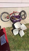 Photo showing red bicycle frame with wheels, purple tote and fan propeller on grass and concrete