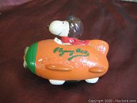 Side view of orange composition coin bank shaped as a plane with Snoopy pilot figure on top and 'Flying Ace' written on side.