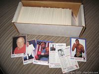 Photo showing a large quantity of boxing cards in a white storage box, with some cards spread out in front including portraits of Cus D'Amato, George Foreman, Joe Louis, Sugar Ray Leonard, and Joe Frazier.