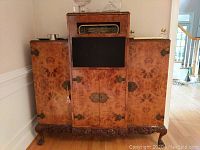 Front view showing full cabinet with burl wood patterns, fabric speaker grille, metal hardware, and carved base.