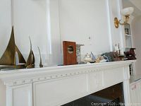 Three brass-colored metal sailboat sculptures on a white mantle with a wooden bookend nearby