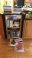 Wide view of kitchen island with multiple stacks of books in cookbooks and gardening genres, and items on floor in front