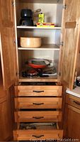 Kitchen cabinet with black fondue pot set on top shelf, wooden bowl, red-orange electric wok with lid in middle shelf, drawers below.