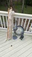 Photo showing the beige knitted outdoor hammock rolled up against white railing, the 18x18 inch outdoor black framed clock leaning against the railing, with the heron and rabbit lawn figurines placed near the clock.
