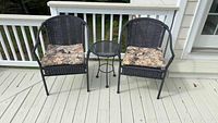Two black wicker outdoor chairs with floral cushion seats with a black metal lattice side table between them on a white deck.