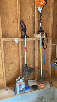Two gas-powered weed trimmers hanging on wall, Stihl and Craftsman, with accessories on ledge beneath.