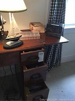 Wide shot of the table lamp, sewing notion boxes and organizer on a wooden desk with drawers beneath and a window with curtains behind.