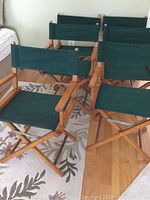 Photo showing six foldable directors chairs with wooden frames and green fabric seats and backs arranged indoors on a hardwood floor with some fabric soiling visible on one chair seat.
