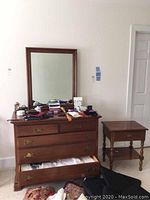 Full view of dresser with mirror mounted and nightstand at side. Dresser top has various clothing items and accessories placed on it.