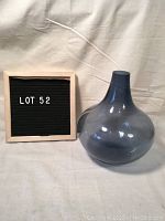 Mid century modern smokey blue ball vase with narrow neck and round body shown next to a sign with lot number