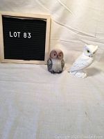 Photo showing both owl figurines side by side on a beige fabric background, identified as lot 83