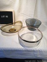 Three glass bowls displayed on white cloth. Left bowl: amber kidney-shaped. Center front: clear with silver rim. Right back: smoky gray large bowl.