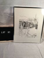 Photo showing framed ink drawing of a village scene with statues and buildings, in silver frame.