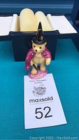 Porcelain bear figurine approximately 3.5 inches tall, wearing a purple cape and black pointed hat, photographed with original box in background and MaxSold auction tag visible.