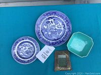 Full view of the lot with two blue Willow pattern plates, one small green hexagonal dish, and one small metal frame on a teal surface.