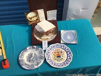 Four collector plates displayed on blue cloth. Includes Quebec Winter plate in brown box with paperwork, blue Jasperware plate in box, and two decorative plates - one with male portrait and one with coat of arms and floral border.