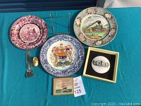 Full lot overview on teal fabric background including 3 plates, framed plate, small painting, gold clog figurine, glass stopper, and plate stand.