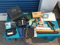 Overview of vintage office supplies lot on a blue table including a black metal cash box, staplers, office tools, paper pads, rulers, and a brass bell