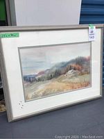 Framed watercolour painting showing rural scene with hills and a house, viewed angled from the left side.