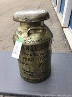 Front view of vintage metal milk can showing handwritten names and numbers all over the body, metal lid in place with hole, side carry handle visible.