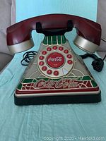 Front view of a Coca Cola styled collectible phone with a rotary dial and handset, showing iconic branding and stained glass effect.