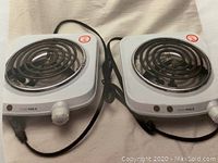 Two white electric hot plate burners with black coil heating elements, control knobs, and power cords on a light background.