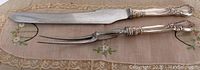Full view of the carving knife and meat fork side by side on lace mat with floral accents.