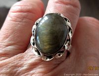 Close-up of oval labradorite cabochon in silver-tone alloy setting on finger, showing the iridescent colors and scalloped bezel.