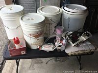 Five large white plastic fermentation buckets with lids and spigots, placed on a table with additional small winemaking accessories and tools around them.