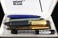 Five vintage fountain pens displayed inside an empty Montblanc Meisterstuck case with white lining and logo on lid.