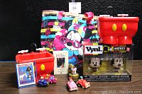Walt Disney collectibles including Mickey and Minnie Mouse Vynl figures, red Mickey Mouse containers, Fantasia VHS tape, Minnie Mouse tote bag, small toy cars, and Swiss Alps puzzle box.