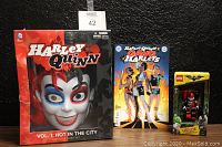 Photo showing Harley Quinn Vol 1: Hot In The City Book with mask set, sealed comic book, and Lego LED Lite Head Lamp figure standing next to each other on black background.