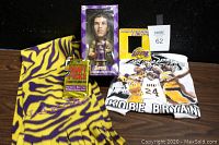 Five Lakers collectibles shown on table including T-shirt, bobble-head, frame, muffler, and card pack.