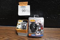 Two brand new old stock Star Wars Darth Vader watches in their original boxes and packaging, one in a metal tin, the other in plastic sealed packaging, photographed against a black background.