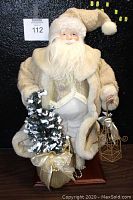 Full view of vintage Santa Claus figurine showing off-white plush coat and holding snow-dusted Christmas tree and decorative lantern on a wooden base.