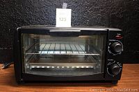 Front view of black toaster oven showing glass door, metal wire rack, metal tray inside, and control knobs on side.