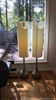 Pair of tall modern floor lamps standing by window with light colored cylindrical fabric shades and brushed metal bases, extra shade not visible in this photo.