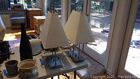 Two contemporary modern silver metal lamps with white pyramidal fabric shades on a white table indoors near a glass door.
