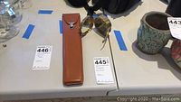 Brown leather case containing ruler next to metal compasses, placed on a table alongside number tags.