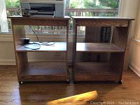 Photo showing two medium-brown wooden computer tables positioned side by side, featuring open shelving and mounted on caster wheels.